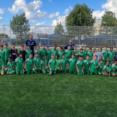 ילדים משחקים כדורגל במגרש מקצועי, לבושים במדי הפועל ובית"ר גדרה, מחייכים ומרימים ידיים בשמחה, על רקע שער ויציעי אצטדיון.